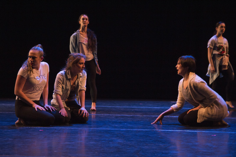 Young girls in rehearsal clothes sit on the floor and stand in the background and appear to be looking for something